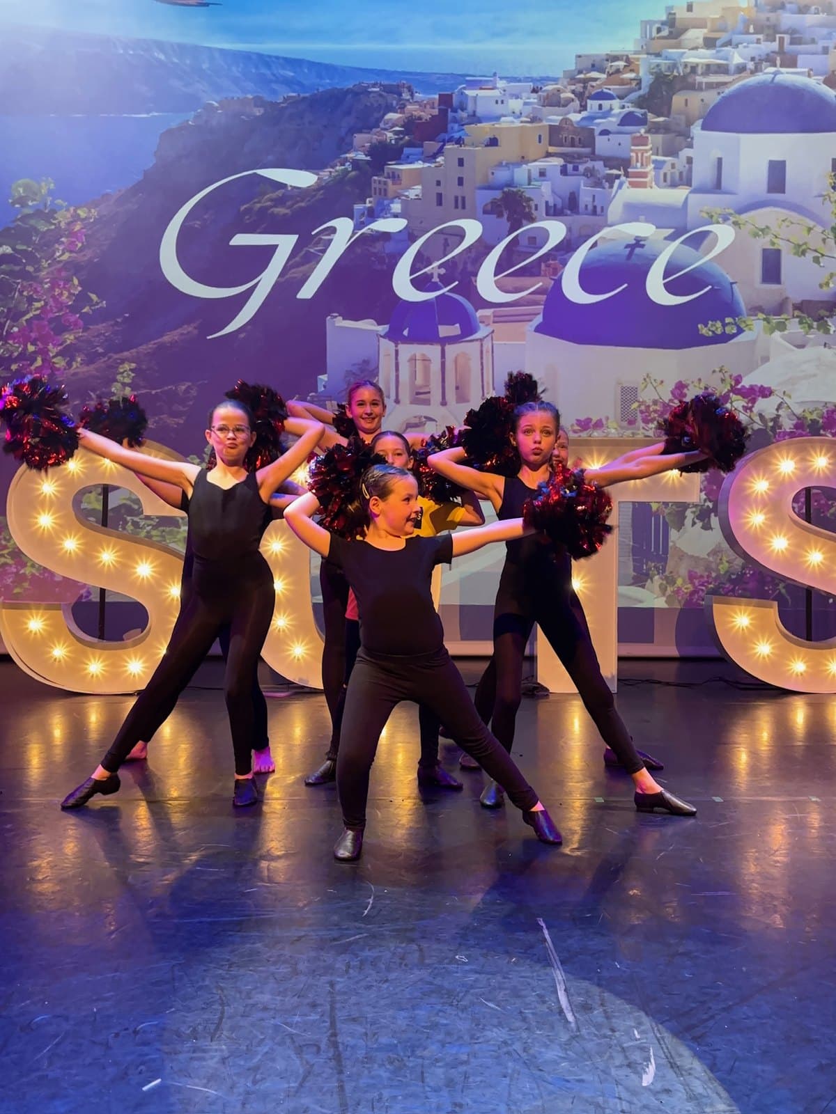 Cheer and pom dancers in black with pom-poms in front of a Greece backdrop